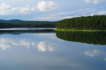 Fototapeta premium Tranquil summer landscape with a forest lake reflecting green trees, a blue sky, and clouds like a mirror