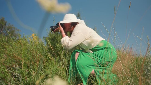 Close up of white wildflower in sharp focus with adult woman blurred in background bending to take photo in grassy field under clear blue sky during sunny day capturing nature