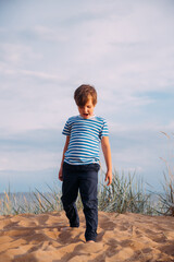 Fototapeta premium Little contented, happy boy on the beach against the background of the sky in the clouds in the grass raising his hands in a sign of success