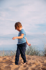 Fototapeta premium Little contented, happy boy on the beach against the background of the sky in the clouds in the grass raising his hands in a sign of success