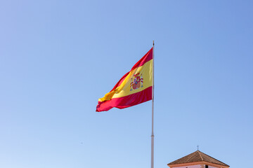 Large Spanish flag waving above the historic cityscape of Cadiz with church towers and old architecture under a clear blue sky. September 2025.