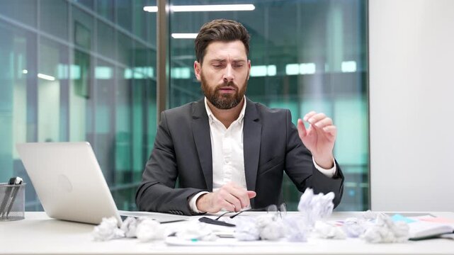 Exhausted businessman is bored sitting at workplace at desk in business office. Overworked upset employee in suit is tired of doing paperwork. Frustrated confused worker has depression, despair