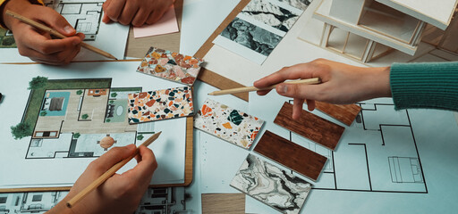 A dynamic scene showcasing hands of individuals engaged in interior design work, analyzing materials and layouts on a table filled with design plans and samples. SACTR