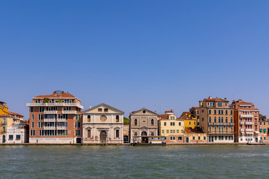 Colorful Venetian canal buildings with church facades. A row of colorful waterfront buildings and two historic church facades along a Venice canal under a bright blue summer sky.