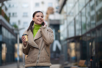 Fototapeta premium Young woman walking on city street holding coffee cup and talking on smartphone, smiling. 