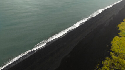 Aerial perspective of a black sand beach meeting the ocean, with white foam waves and vibrant green vegetation contrasting the volcanic terrain.