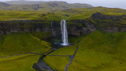 Seljalandsfoss waterfall cascades from a tall cliff into a pool, surrounded by moss covered hills, walking paths, and smaller distant waterfalls.