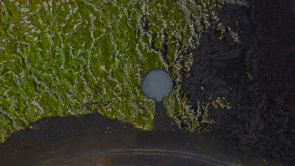 Aerial perspective of a small circular water body surrounded by green moss within a dark volcanic terrain, showcasing Iceland's dramatic geology.