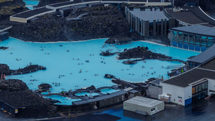 The Blue Lagoon's milky blue waters contrast with dark lava fields, featuring visitors, modern spa facilities, and rugged volcanic surroundings. © Ekaterina