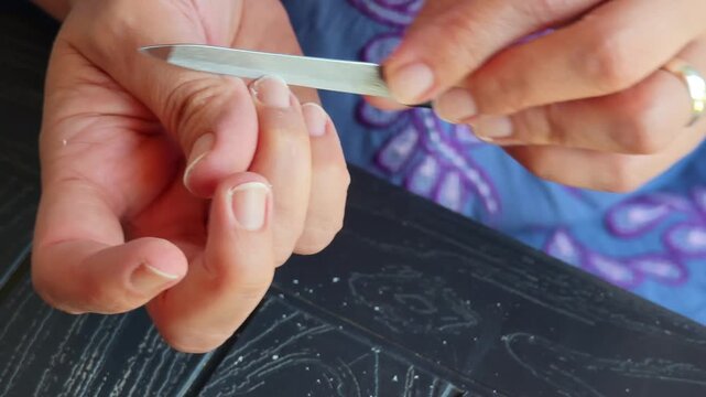 Woman carefully filing and shaping fingernails with metal nail file during home beauty routine, showcasing precise nail care and personal grooming process
