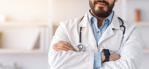 Modern telemedicine, health care, therapist and doctor advice during lockdown. Serious confident attractive arabian guy in white coat, crossed arms on chest in office interior in clinic, free space