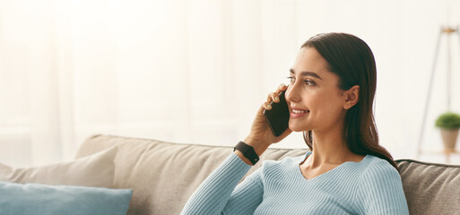 Female speaking on mobile phone with client, use laptop for remote work at home, new normal, workspace at covid-19 lockdown. Cheerful young arab female on sofa in living room interior make call