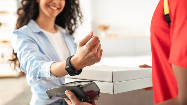 Portrait of smiling woman making payment through wireless modern bank terminal. Consumer paying using smart watch and NFC technology to courier for package delivery. Modern Futuristic Technology