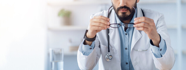Problems at work, overwork in clinic, first symptoms of disease. Tired sad millennial arabian bearded guy doctor in white coat takes off his glasses, sits at desk in office interior, free space