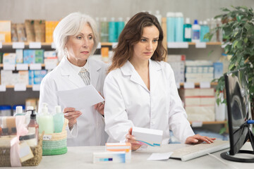 Fototapeta premium Woman pharmacist works at computer, intern observes actions of professional, asks questions, practices work skills. On line document flow, checking drug instruction
