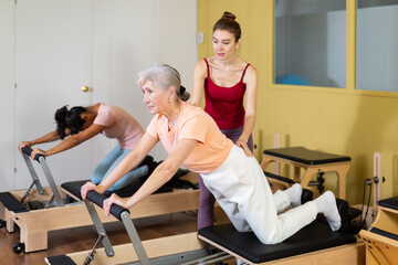 Friendly young girl professional pilates instructor assisting elderly woman to do exercises on...