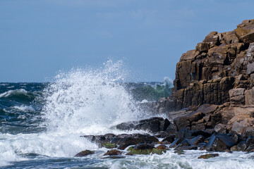 Waves crash against a rocky coastline, sea spray and whitewater under blue sky