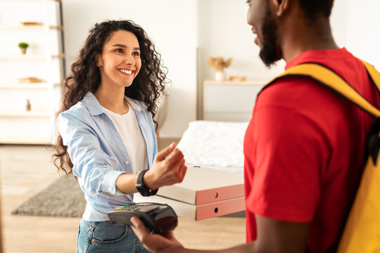 Delivery And Technology Concept. Smiling black male courier in red uniform and cap holding POS machine in hand and pizza boxes, woman using smart watch for contactless payment, standing at doorstep