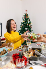 Young woman laughing while receiving a bowl of salad during christmas eve dinner celebration with...