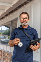 A home inspector with an iPad holding a water pressure meter in his hand, looking at the camera, smiling