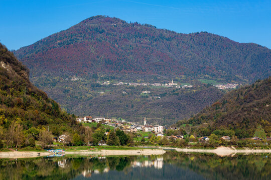 Scenic landscape featuring a serene lake Lago di Corlo reflecting a vibrant mountain range with lush greenery and a quaint village nestled at the foot, showcasing natural beauty and tranquility