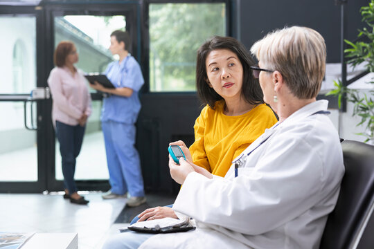 Medical assistant conducting blood sugar levels measurement with glucometer during routine medical exam in clinic lobby. Asian patient discussing glucose readings and preventive care steps.