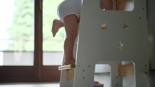 Close-up of a baby climbing a white wooden stepping stool in a kitchen, showing legs and feet, conveying a milestone in independence and curiosity