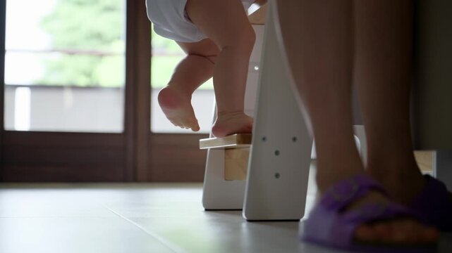 Low angle shot of a baby climbing a wooden stepping stool with mom or dad standing nearby, showing a milestone in childhood learning and independence in the kitchen