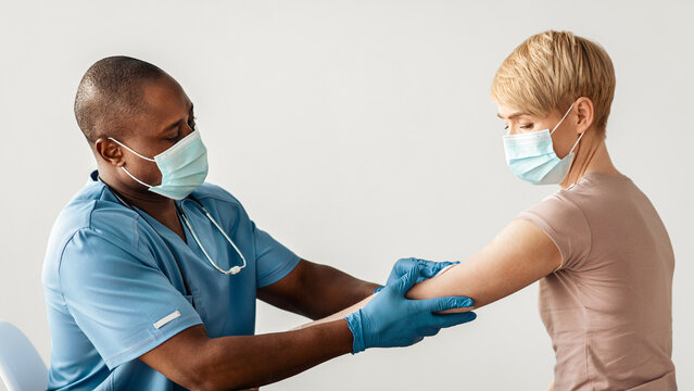 Health care, vaccination of population and modern treatment against covid-19 epidemic. Adult african american doctor or nurse in protective mask prepares hand for injection for mature woman in clinic - Powered by Adobe