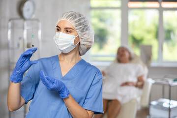 Obraz premium Portrait of young nurse wearing blue medical scrubs, facial mask and disposable gloves with syringe in her hands