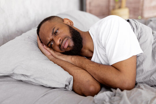 Depression Concept. Portrait of upset African American man lying in bed in the morning and thinking. Sad black male feeling lonely or stressed, suffering from lack of sleep, distraught - Powered by Adobe