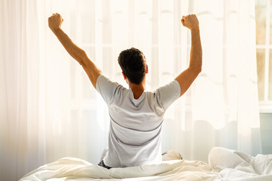 Good morning. Young man waking up and stretching his arms in cozy bedroom, back view. Guy sitting on bed facing the window, awakening after good sleep and rest - Powered by Adobe