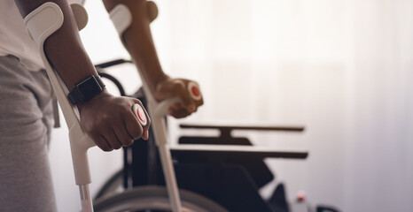 Fototapeta premium Physical activity and training to recover from injury and illness. Adult african american man disabled with crutches walks near wheelchair on large window background in interior, cropped, close up