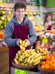 Positive male seller in apron displaying assortment of bananas at supermarket