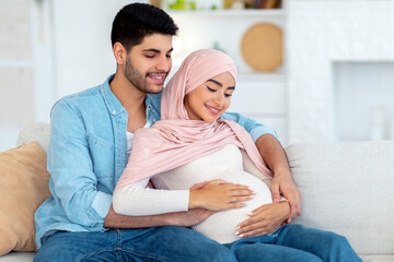 Fototapeta premium Loving expecting arab couple cuddling while spending time together at home, sitting on sofa, free space. Pregnant muslim couple hugging wife's big belly and smiling