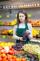 Young positive female seller in apron displaying assortment of grape at supermarket