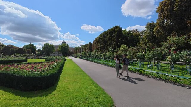 Młoda para spacerująca alejką w słoneczny, letni dzień w parku Volksgarten w Wiedniu, Austria. Wok&oacute;ł widać pięknie utrzymane ogrody r&oacute;żane, zielone trawniki i charakterystyczne parkowe krzesła. W tle 