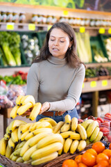 Young woman shopper in casual clothes chooses bananas in vegetable shop