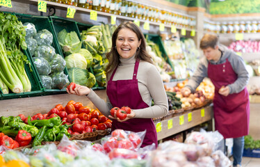 Smiling young saleswoman taking tomatoes from food stall in large grocery store