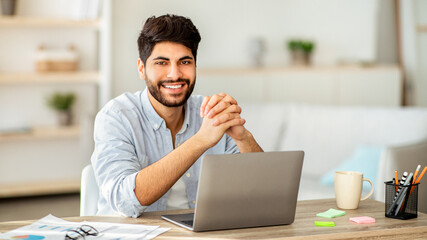 Obraz premium Successful freelancer. Happy arab man sitting at desk with laptop and smiling to camera while working witn documents on computer at home. Freelance career, remote job
