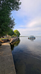 Fototapeta premium A serene lakeside view with a calm water surface reflecting the sky. A boat is anchored near the shore, surrounded by green trees and a concrete walkway.