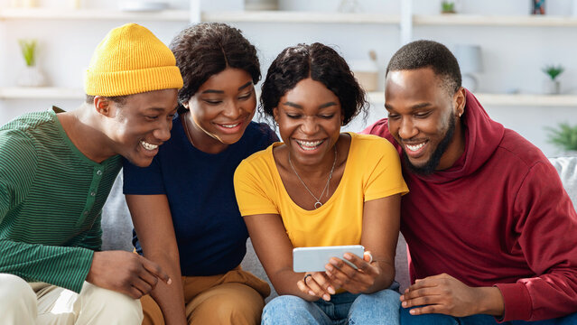 Happy african american fellows sitting on couch at home, using mobile phone together, laughing, having video conference with friends. Positive black men and women watching funny videos on smartphone