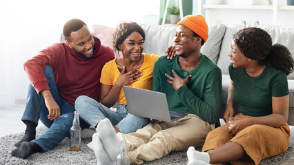 Happy black friends young men and women using laptop, sitting on floor in living room, watching movie or video online and laughing, african american couples having video conference at home