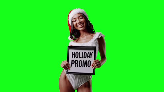 Realistic video of a smiling model with dark skin and long wavy black hair wearing a white Christmas outfit with a Santa hat, holding a sign that says &ldquo;Holiday Promo&rdquo;, isolated on a bright green chrom