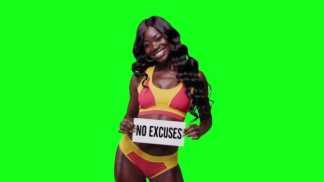 Realistic video of a happy fitness model with dark skin and long wavy black hair wearing a pink sports bra, holding a sign that says &ldquo;No Excuses&rdquo;, smiling at the camera and isolated on a bright green 
