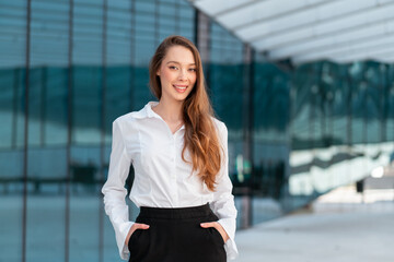Confident businesswoman wearing white shirt and black pants standing with hands in pockets near glass building, smiling and reflecting modern professionalism and charm