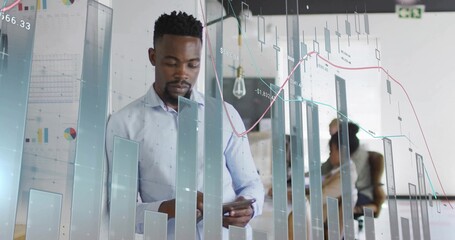 Analyzing business-casual man viewing touchscreen device in open-plan office, with financial charts