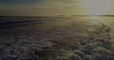 Fototapeta premium Rolling foamy ocean waves catching golden sunset light at beach viewpoint, coastal seascape