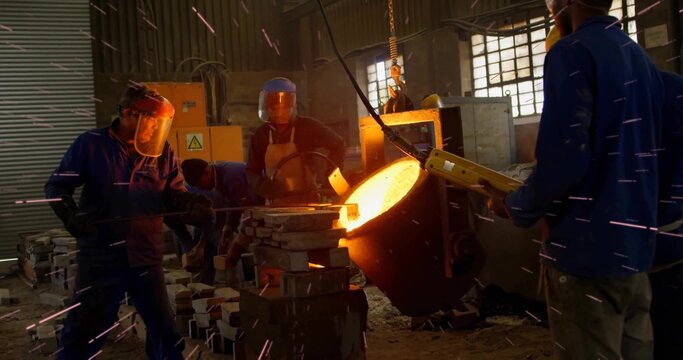Pouring workers guiding molten metal from ladle in foundry shop with hoist chain molds face shields
