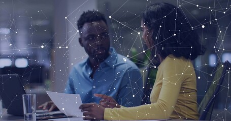 Yellow-top woman holding document guiding man typing on laptop at office desk, with network overlay